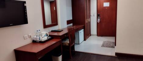 A hotel room with a wooden desk, a flat-screen TV, a mirror, a mini fridge, and a small seating area.