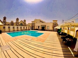 Una piscina con solárium de baldosas, rodeada de edificios con ventanas de arco y una torre del reloj.