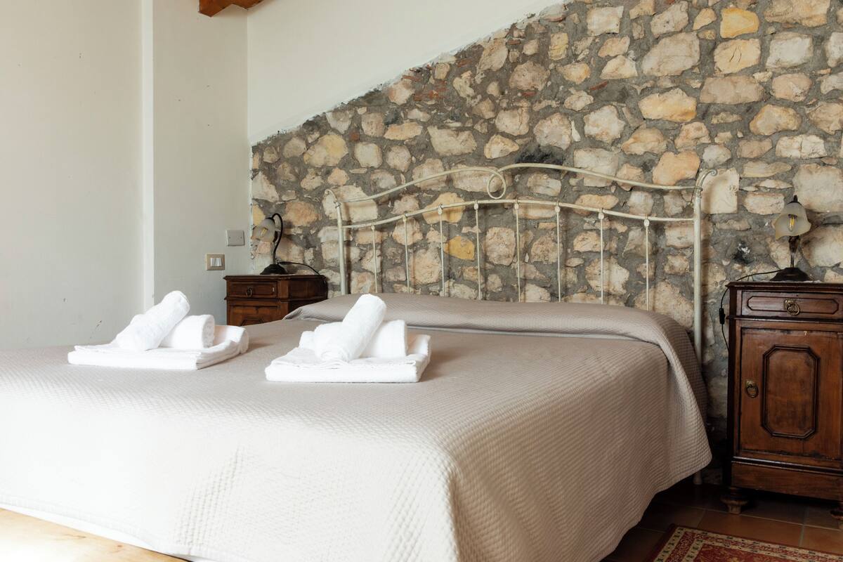 A bedroom with a stone wall, a metal bed frame, a wooden nightstand, and a nightstand lamp.