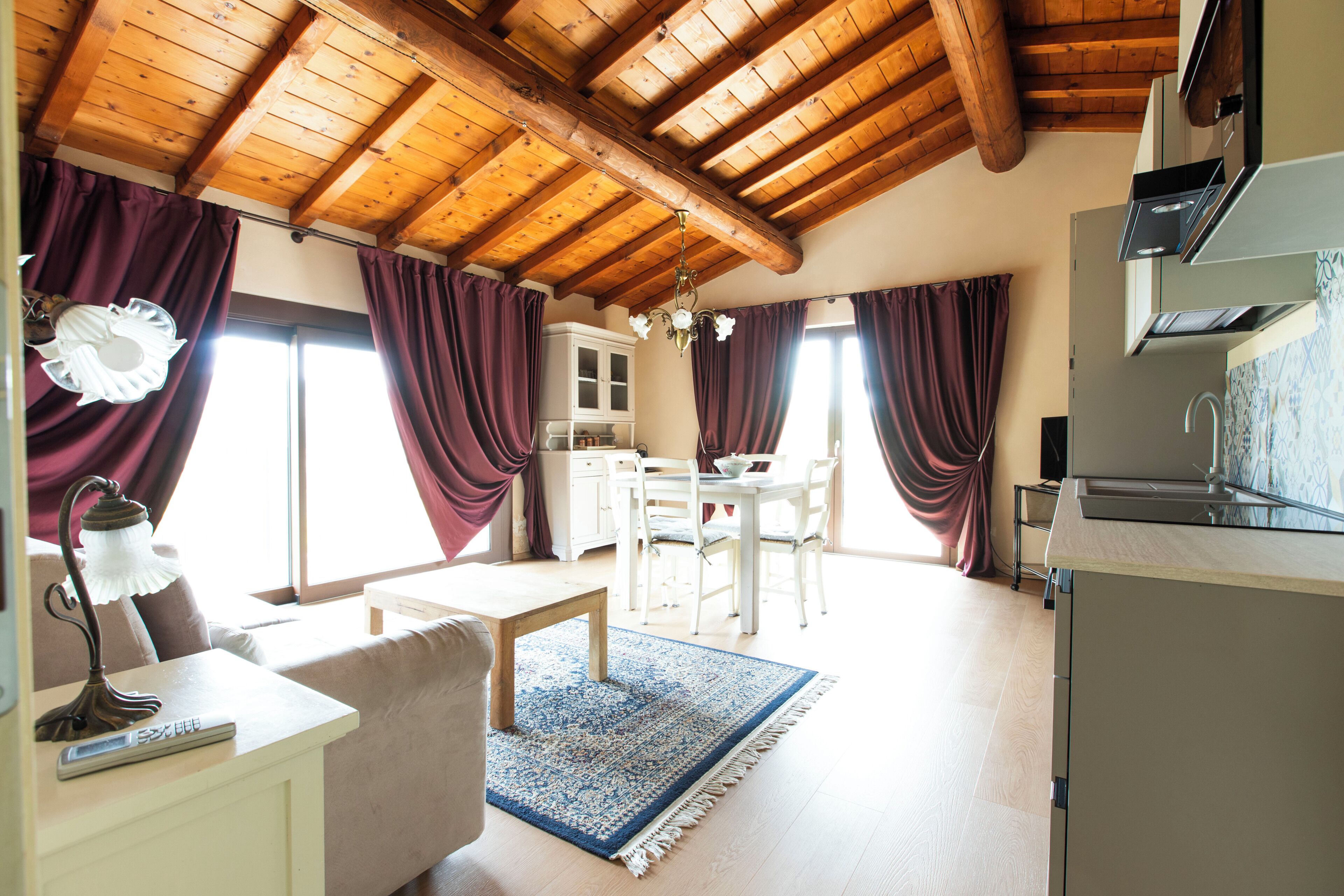 Ein Zimmer mit Holzbalkendecke, einem Esstisch mit Stühlen, einer Küchenzeile und einem Fenster mit Vorhängen.
