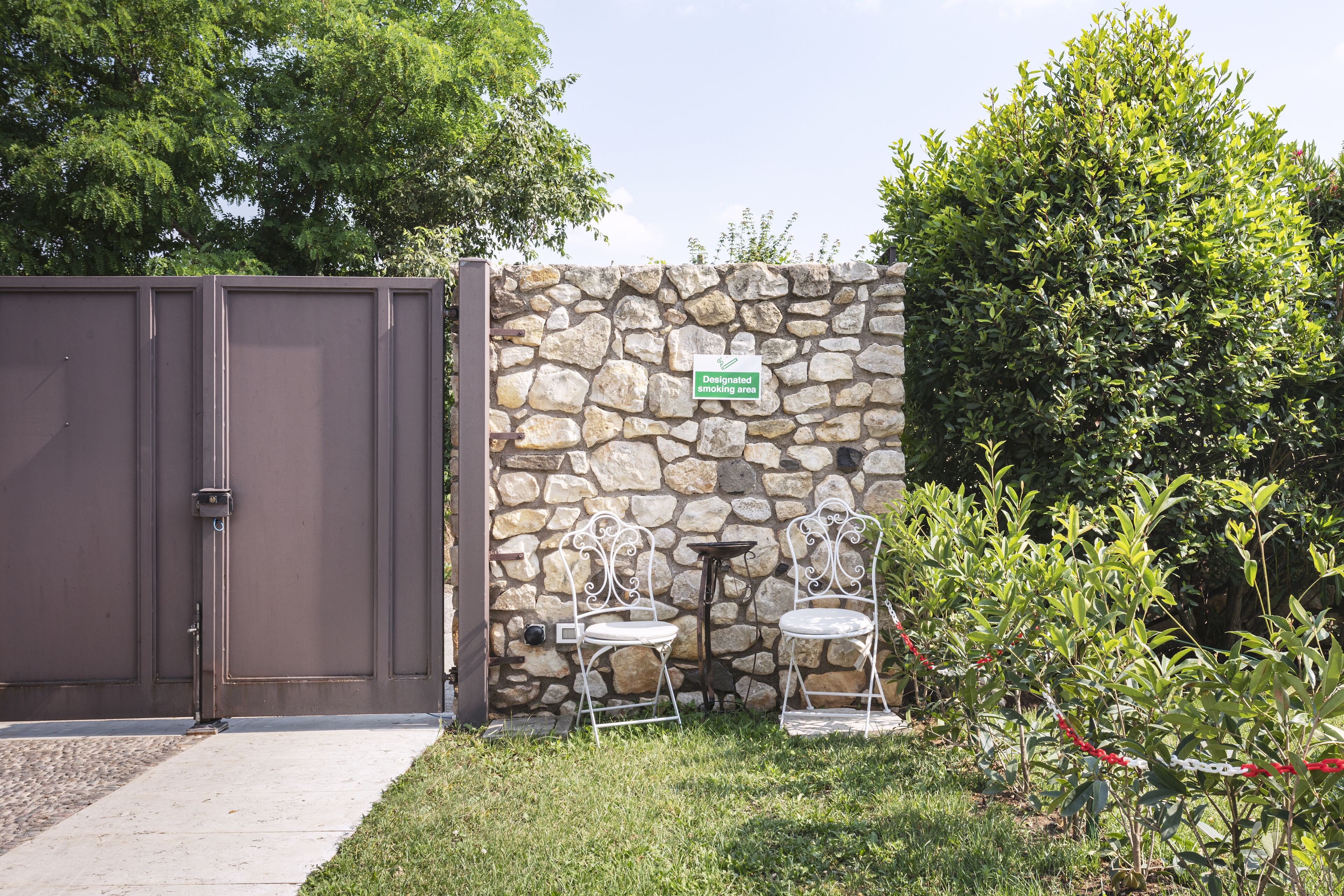 Un muro de piedra con un cartel de zona para fumadores, una reja de metal y dos sillas de metal blanco en un área con césped.