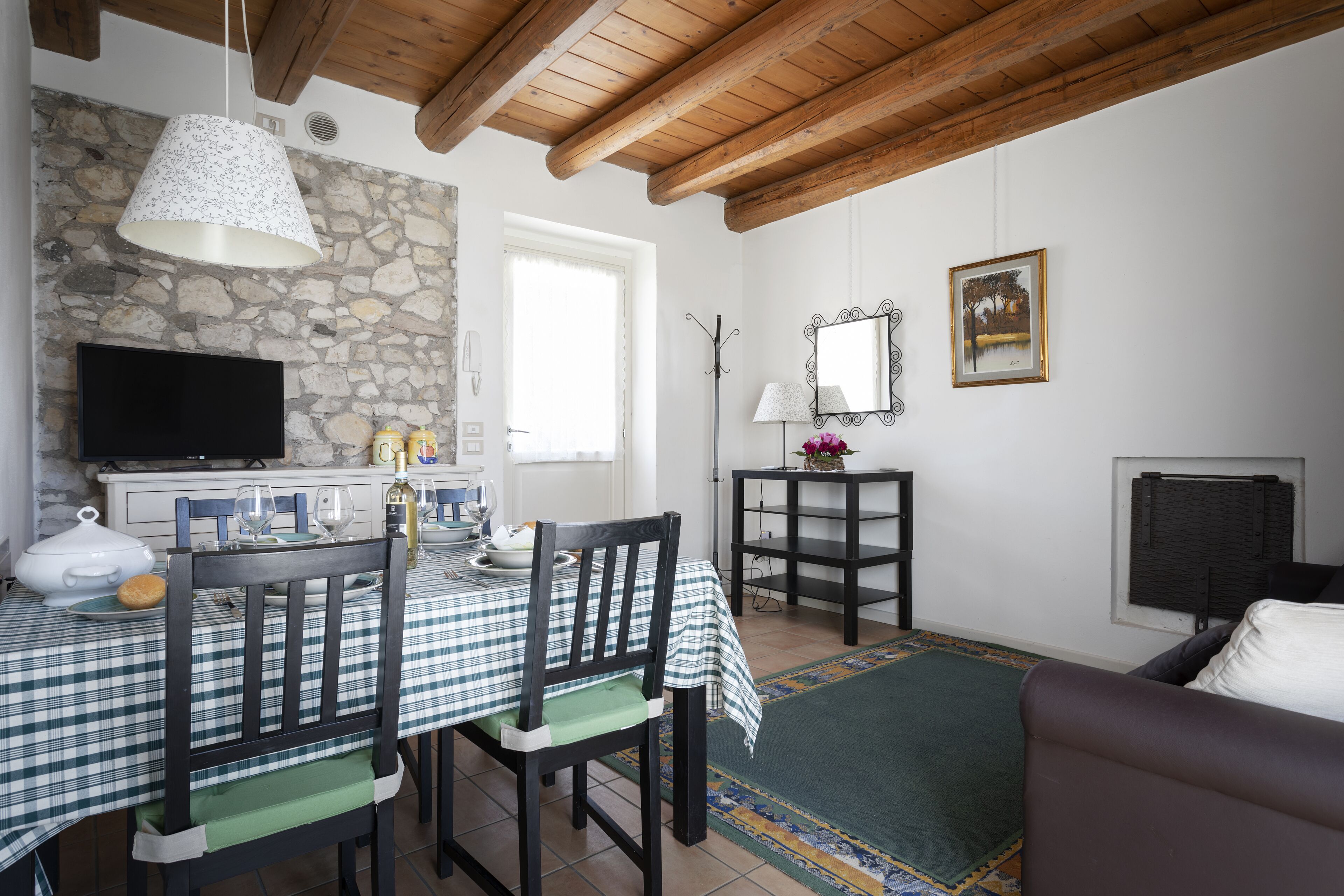A dining area with a checkered tablecloth, wooden chairs, a television, stone wall, and a fireplace.