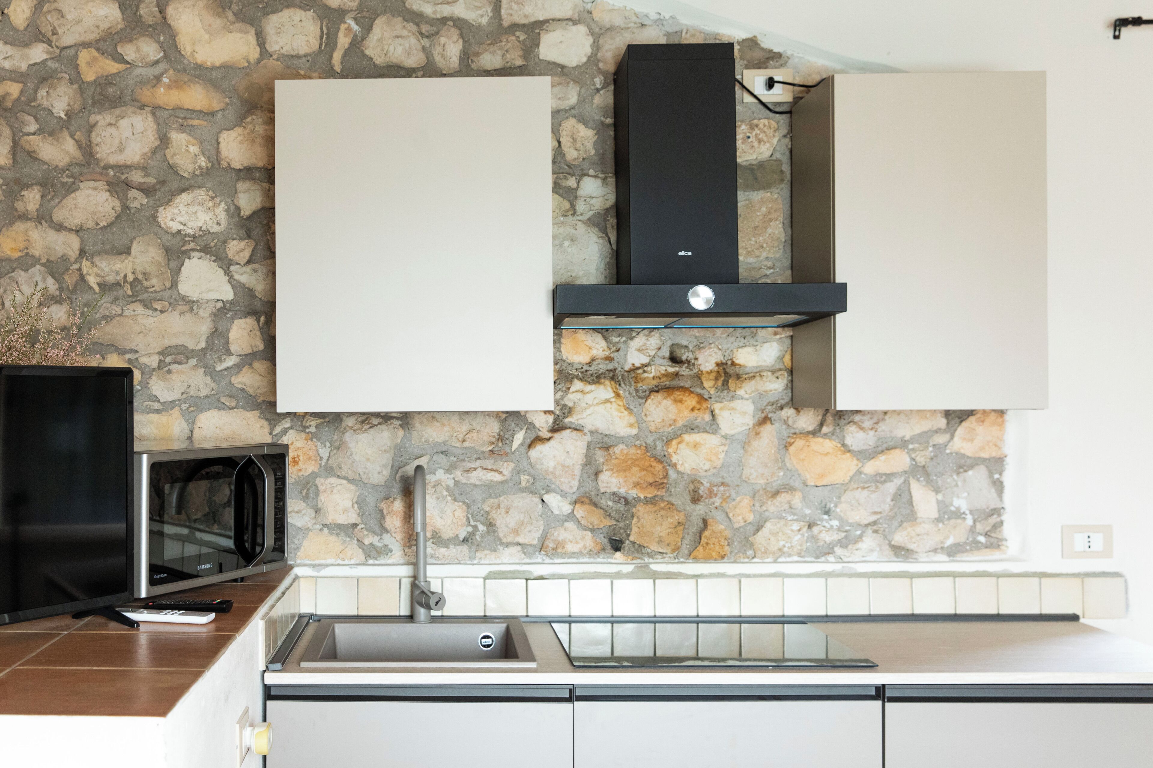 A kitchen with a stone wall, a microwave, a sink, and cabinets.