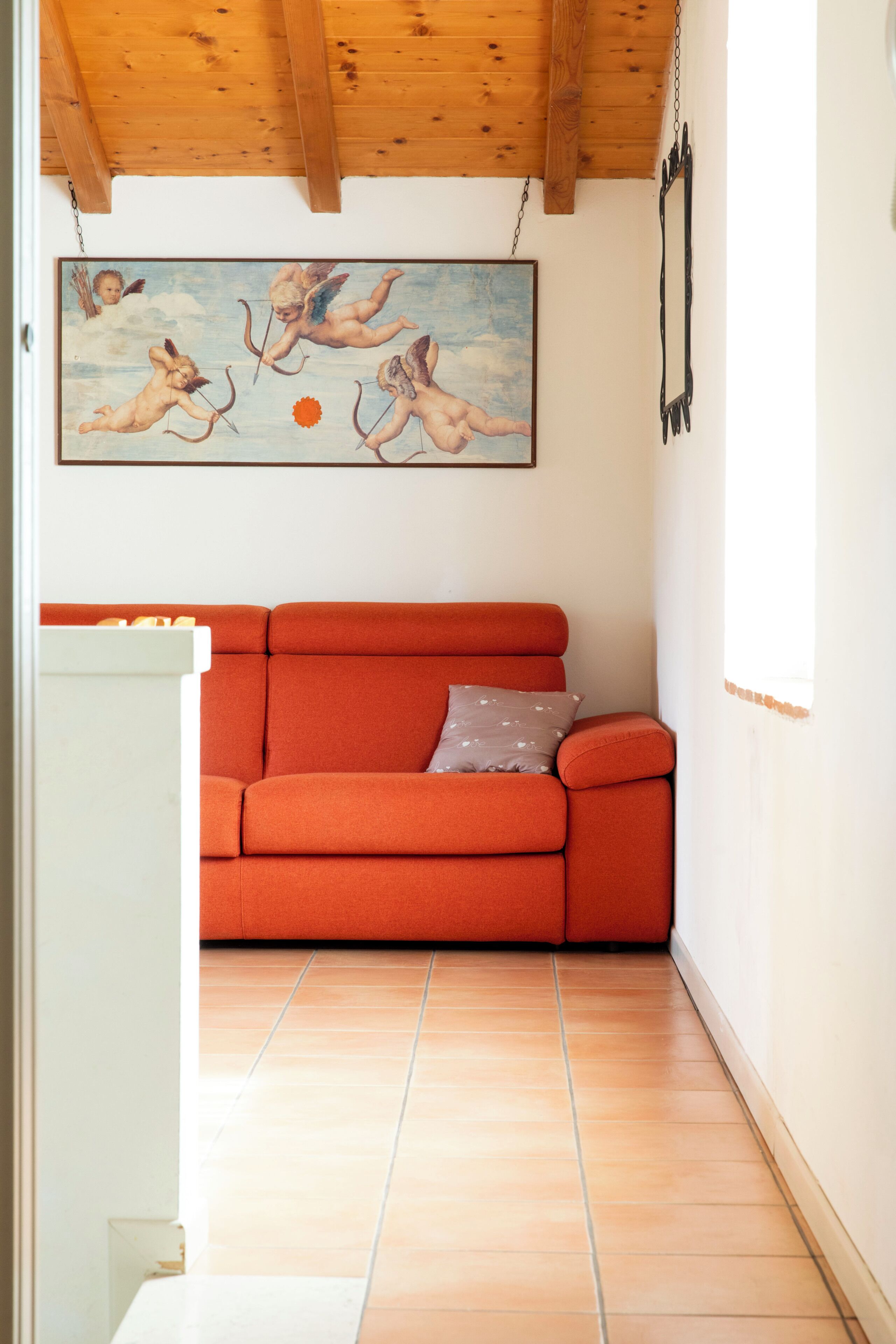 Una habitación con techo de madera, un sofá rojo, un cuadro de querubines y piso de baldosas.