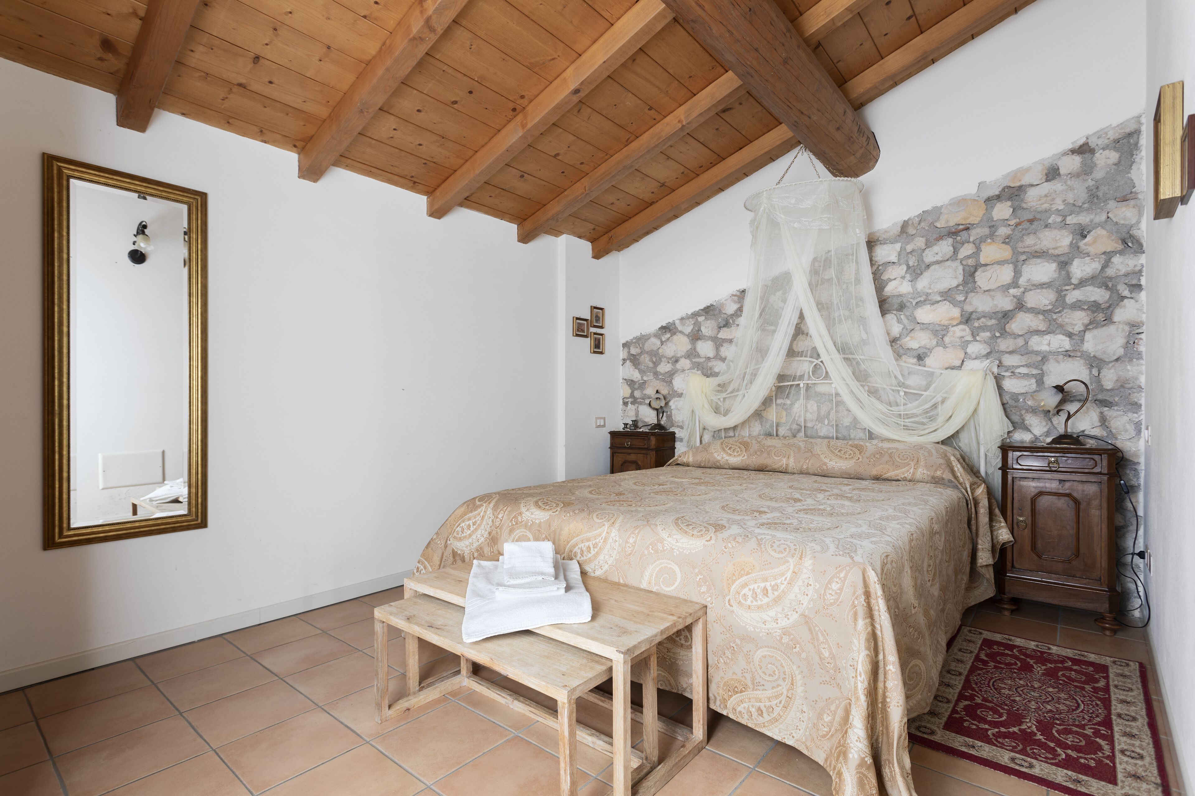 Un dormitorio con techo de madera, pared de piedra, cama con dosel, mesita de noche con toalla y otra mesita de noche con lámpara.