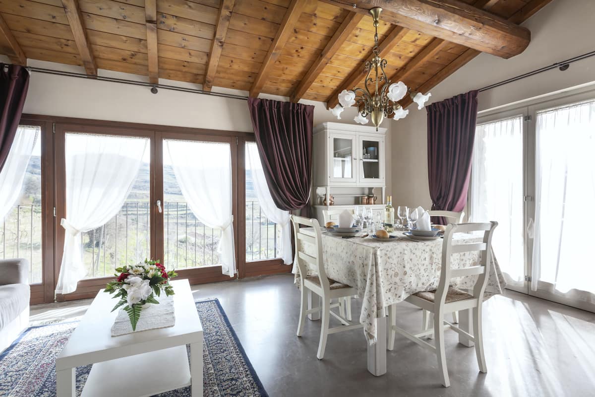 A dining area with a wooden ceiling, white table, and chairs. There is a window with curtains and a view of the outdoors.