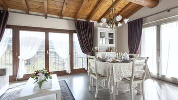 Un comedor con techo de madera, mesa y sillas blancas. Se ve una ventana con cortinas y vistas al exterior.