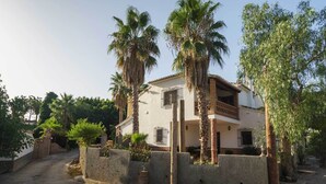 Une maison à deux étages avec un balcon, entourée de palmiers et d’un mur de pierre.