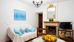 Un salon chaleureux avec un canapé blanc, des coussins bleus, une cheminée, un téléviseur et un tableau accroché au mur.