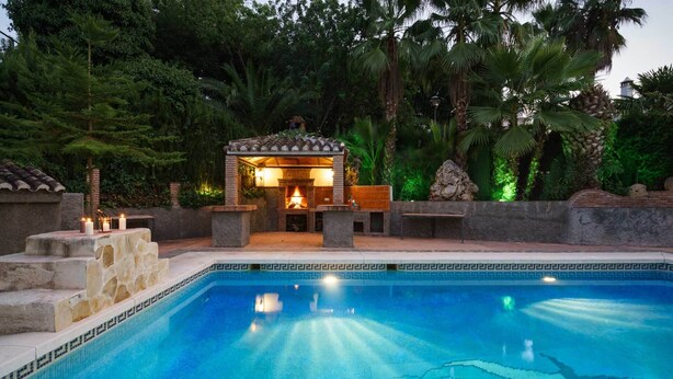 Une piscine bordée de pierres, un espace aménagé avec un foyer extérieur et une végétation luxuriante tout autour.