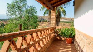 Un balcon doté d’une balustrade en bois, d’une plante en pot et offrant une vue sur un paysage vallonné, avec des arbres et un ciel dégagé.