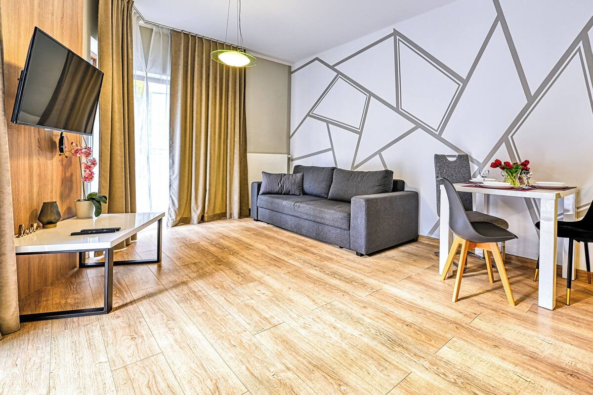 Una sala moderna con un sofá, una mesa de comedor con sillas, un televisor de pantalla plana montado en la pared y piso de madera.