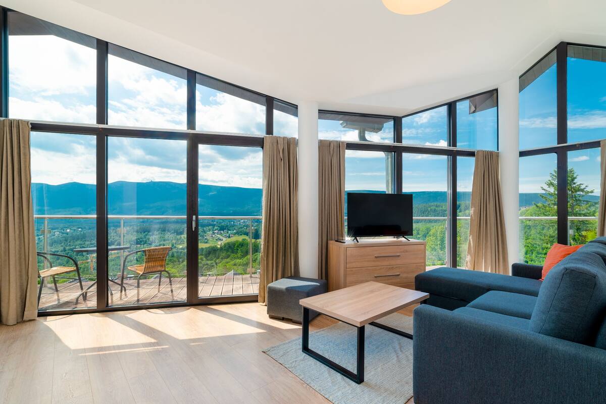 Amplia sala de estar con ventanas grandes que ofrecen vistas a la montaña, un televisor de pantalla plana sobre un mueble de madera y un sofá confortable.
