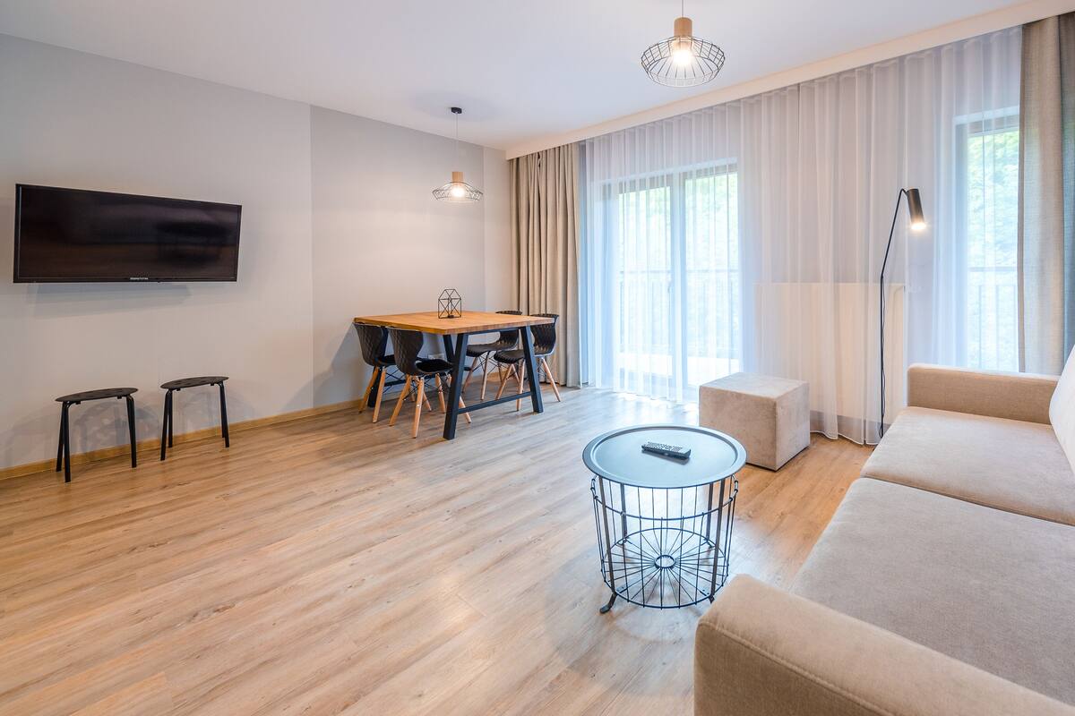 Una sala de estar moderna con un sofá, una mesa de comedor con sillas, un televisor montado en la pared y un amplio ventanal con cortinas.