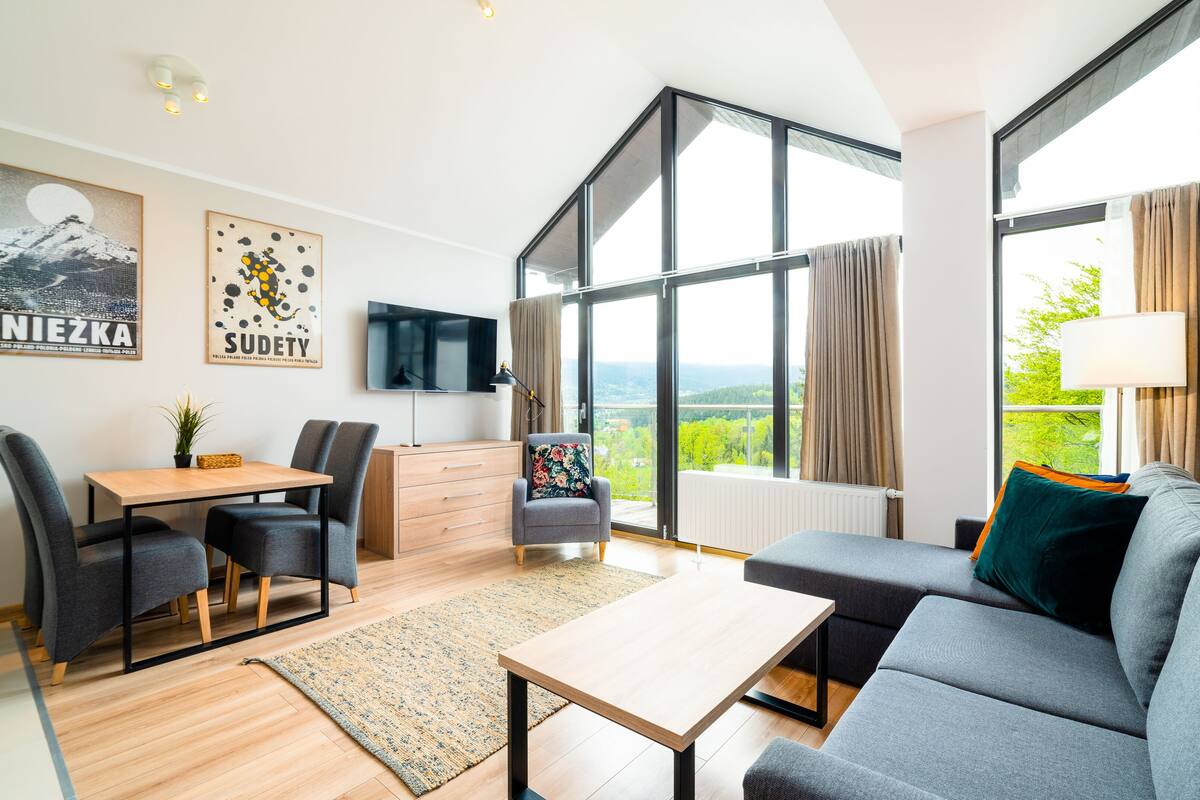 Una sala moderna con sofá, mesa de comedor y sillas. Hay un televisor montado en la pared y ventanas grandes con cortinas.