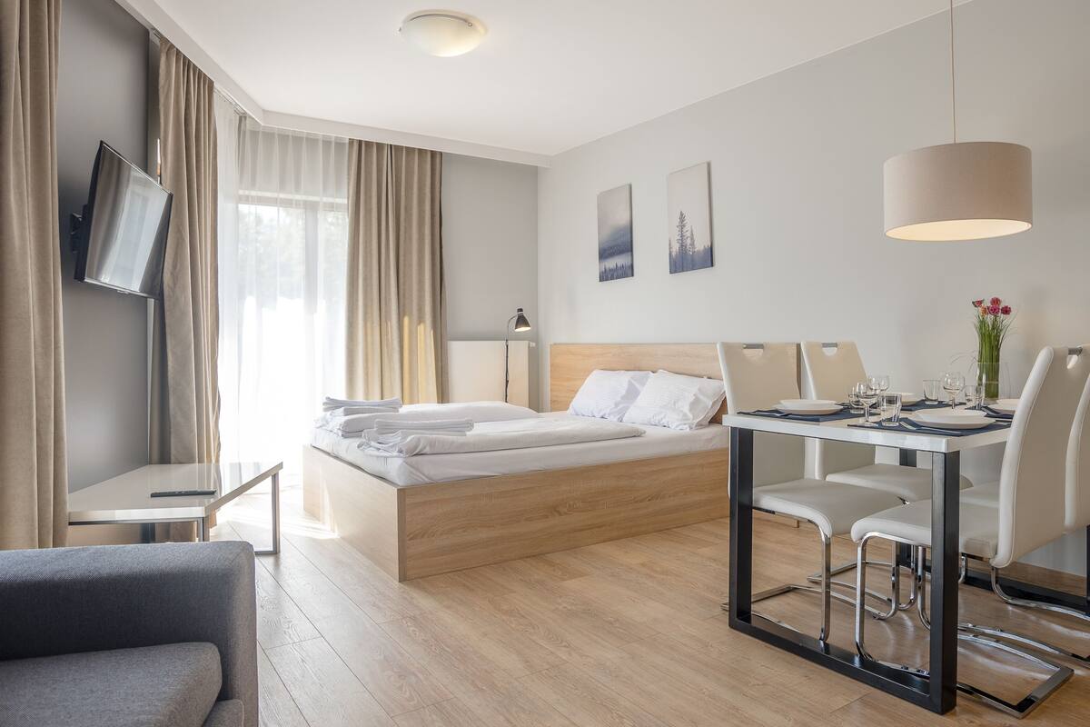 Una habitación de hotel moderna con una cama, una mesa de comedor con sillas, un televisor montado en la pared y una lámpara en el techo.