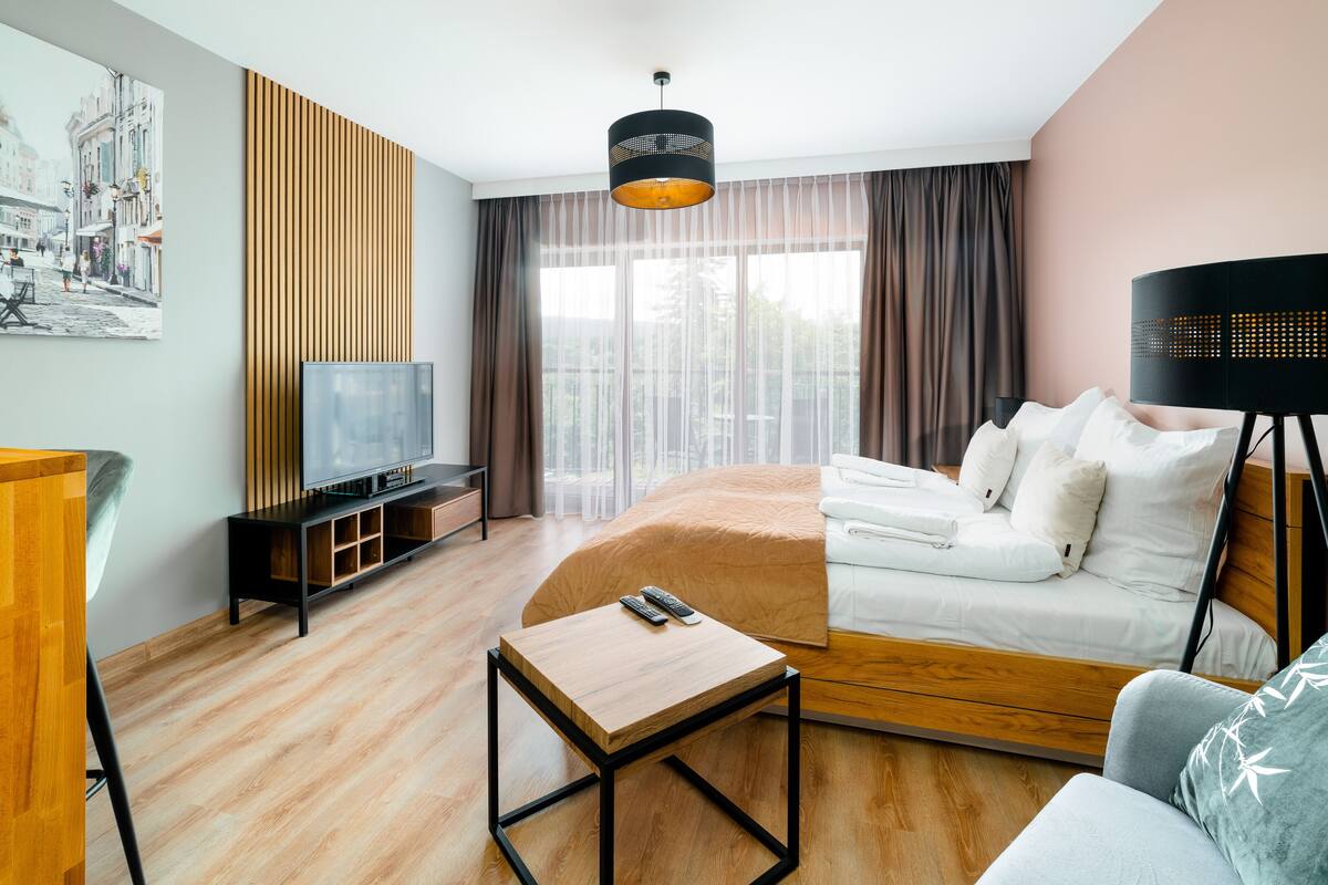 Una habitación de hotel moderna con suelo de madera, una cama con ropa blanca, un sofá, una mesita, un televisor sobre un mueble de madera y un ventanal con cortinas.