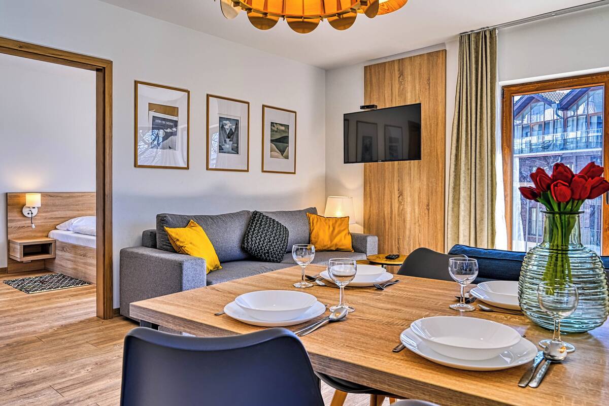 Una habitación de hotel moderna con comedor, sofá, televisión montada en la pared y ventana con vistas a edificios.