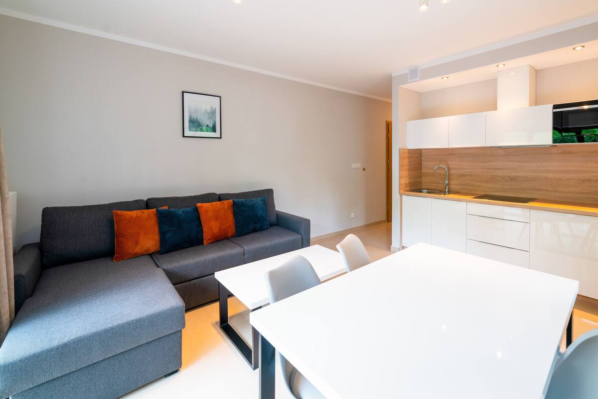 Una sala moderna con un sofá gris, una mesa de comedor blanca y una zona de cocina con encimera de madera y un hervidor.