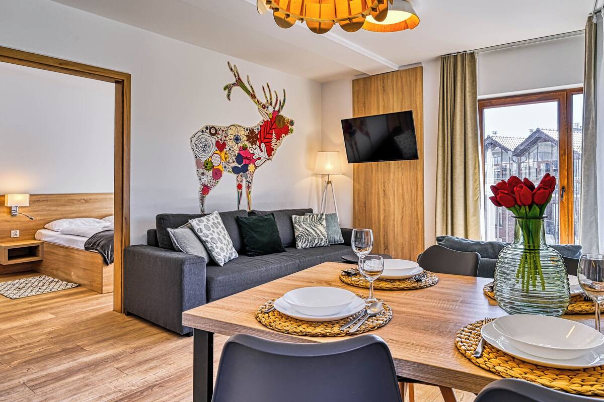 Una habitación de hotel moderna con piso de madera, un sofá gris con cojines estampados, una mesa de comedor dispuesta con platos y vasos, un florero con flores rojas, y un televisor montado en la pared.