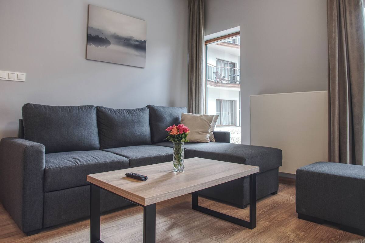 Una sala de estar moderna con un sofá gris, una mesa de centro de madera y un pequeño taburete. Un jarrón con flores está sobre la mesa.