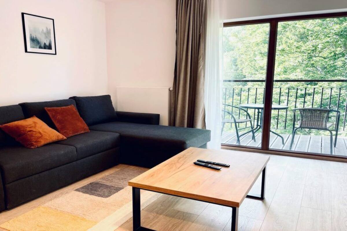 Una sala moderna con un sof á oscuro, dos cojines, una mesa de centro de madera y un amplio ventanal corredero que da a un balcón exterior con sillas.