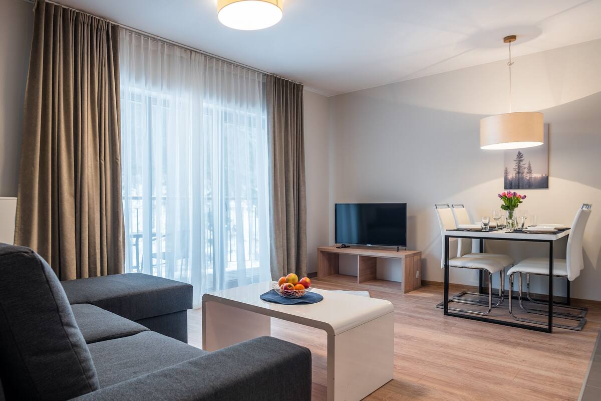 Una habitación de hotel moderna con sofá, una mesita con fruta, televisión, escritorio con sillas y un cuadro en la pared.