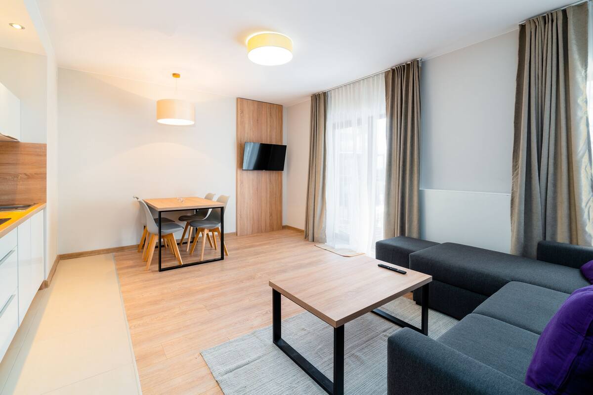 Una sala de estar moderna con un sofá, una mesa de centro, un televisor montado en la pared y una zona de cocina con un lavamanos y armarios de madera.
