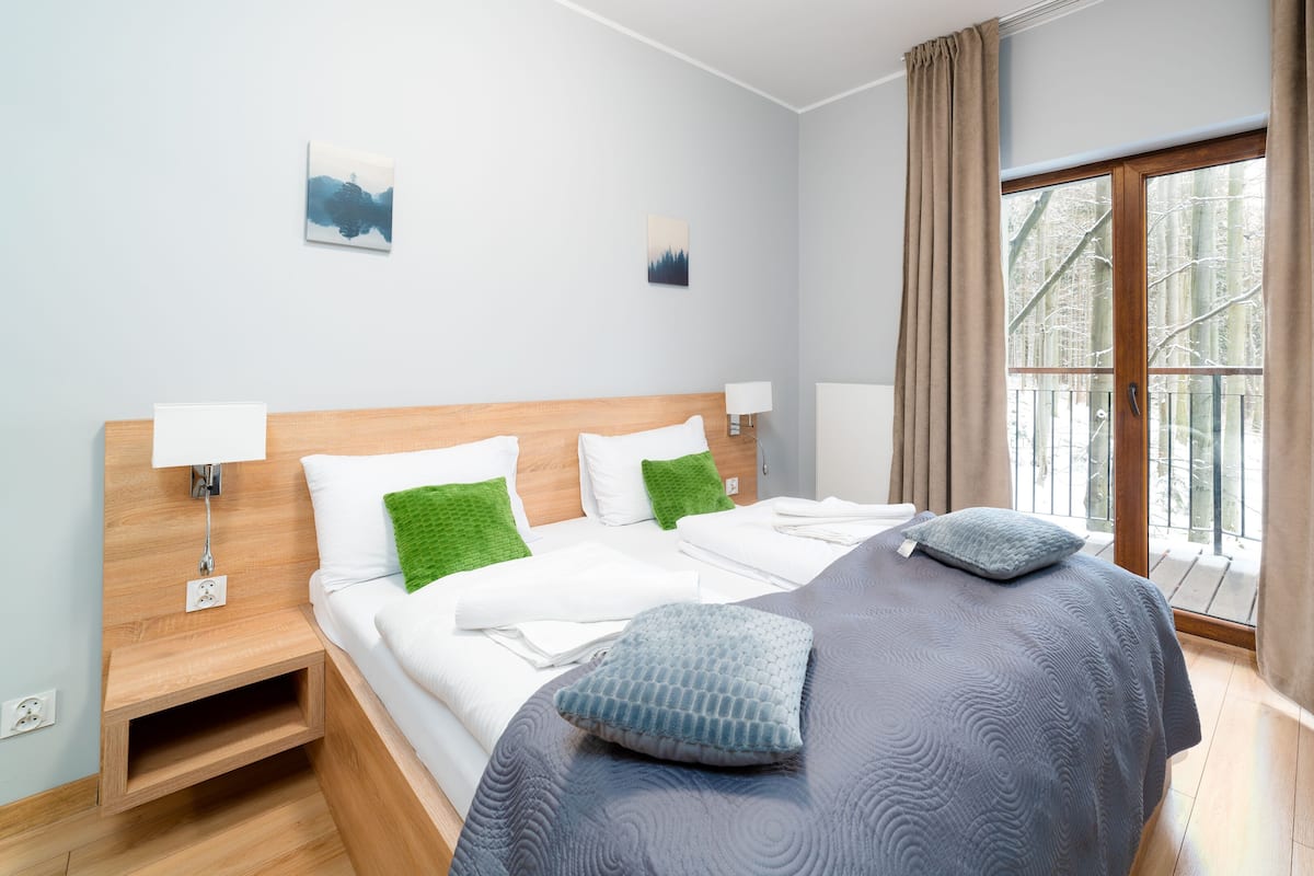 Habitación de hotel con cama de madera, ropa de cama blanca, almohada verde, manta gris, mesita de noche con lámpara y ventana con cortinas.