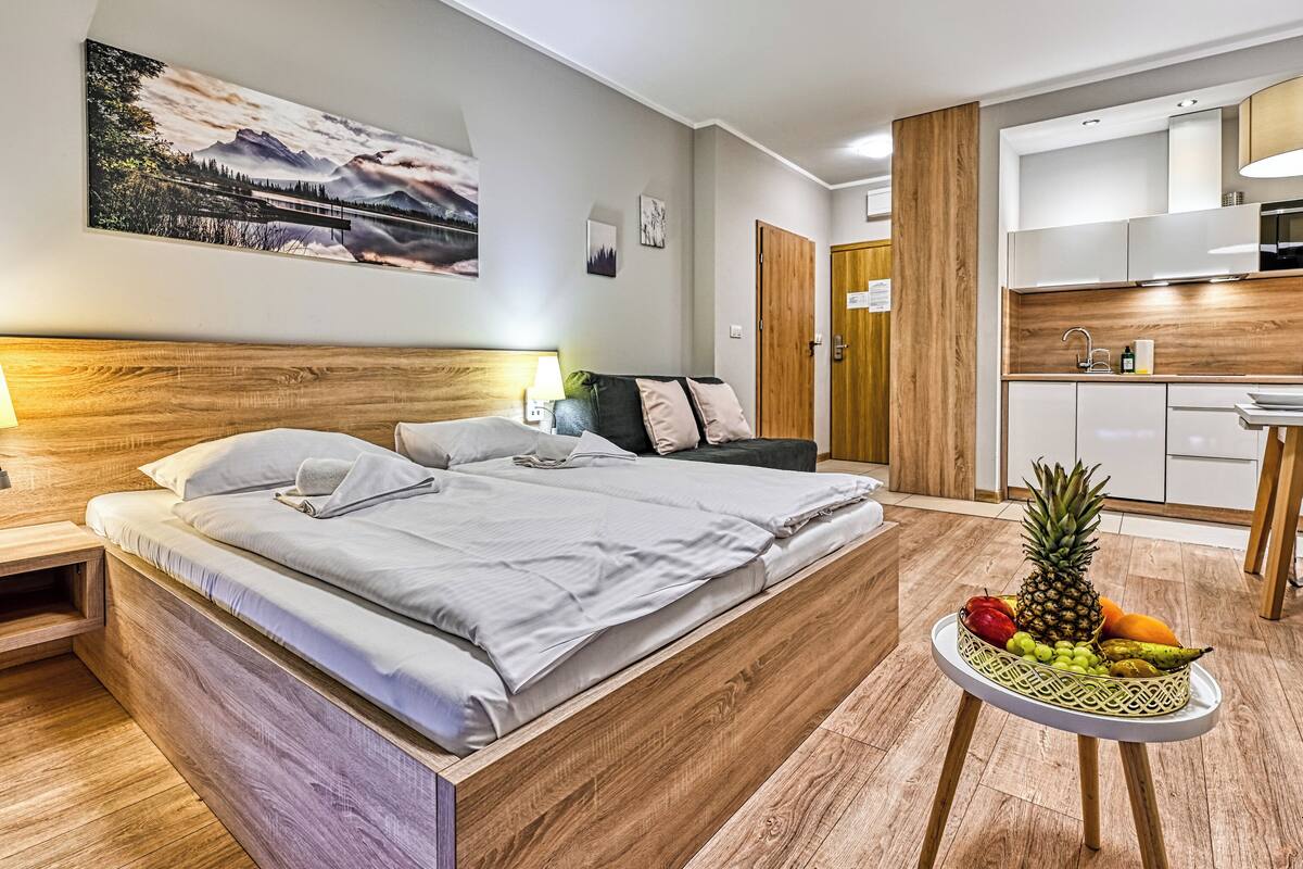 Una habitación de hotel moderna con cabecera de madera, cama con ropa de cama blanca, mesita de noche con lámpara, una pequeña mesa redonda con fruta y una kitchenette con microondas y fregadero.