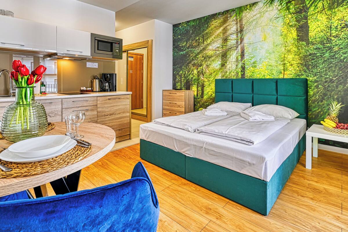 Una habitación de hotel moderna con cama, una mesa de comedor con sillas, una zona de cocina y un gran mural con motivos forestales.