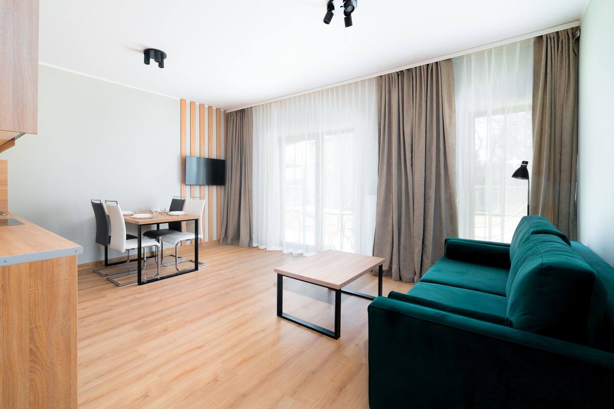 Una sala moderna con un sofá verde, una mesa de comedor de madera con sillas, un televisor de pantalla plana en la pared y una zona de cocina con hervidor.