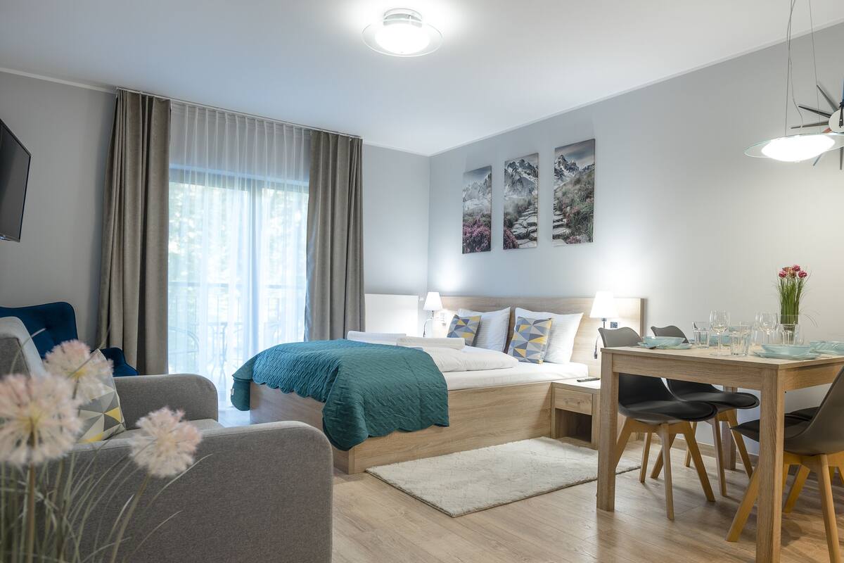 Una habitación de hotel moderna con cama, mesita de noche y zona de comedor con sillas y una mesa.