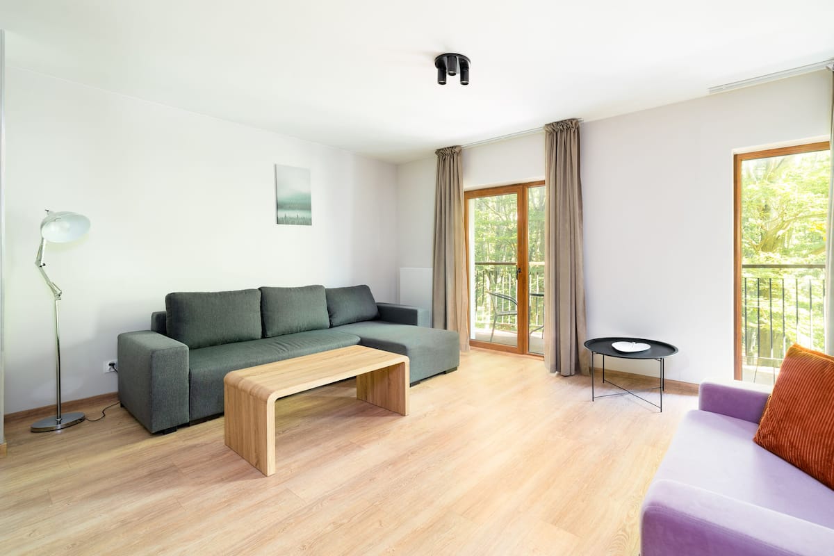 Una sala moderna con un sofá gris, una mesa de centro de madera, un sofá púrpura con un cojín rojo, una lámpara de pie, un televisor montado en la pared y un balcón con cortinas.