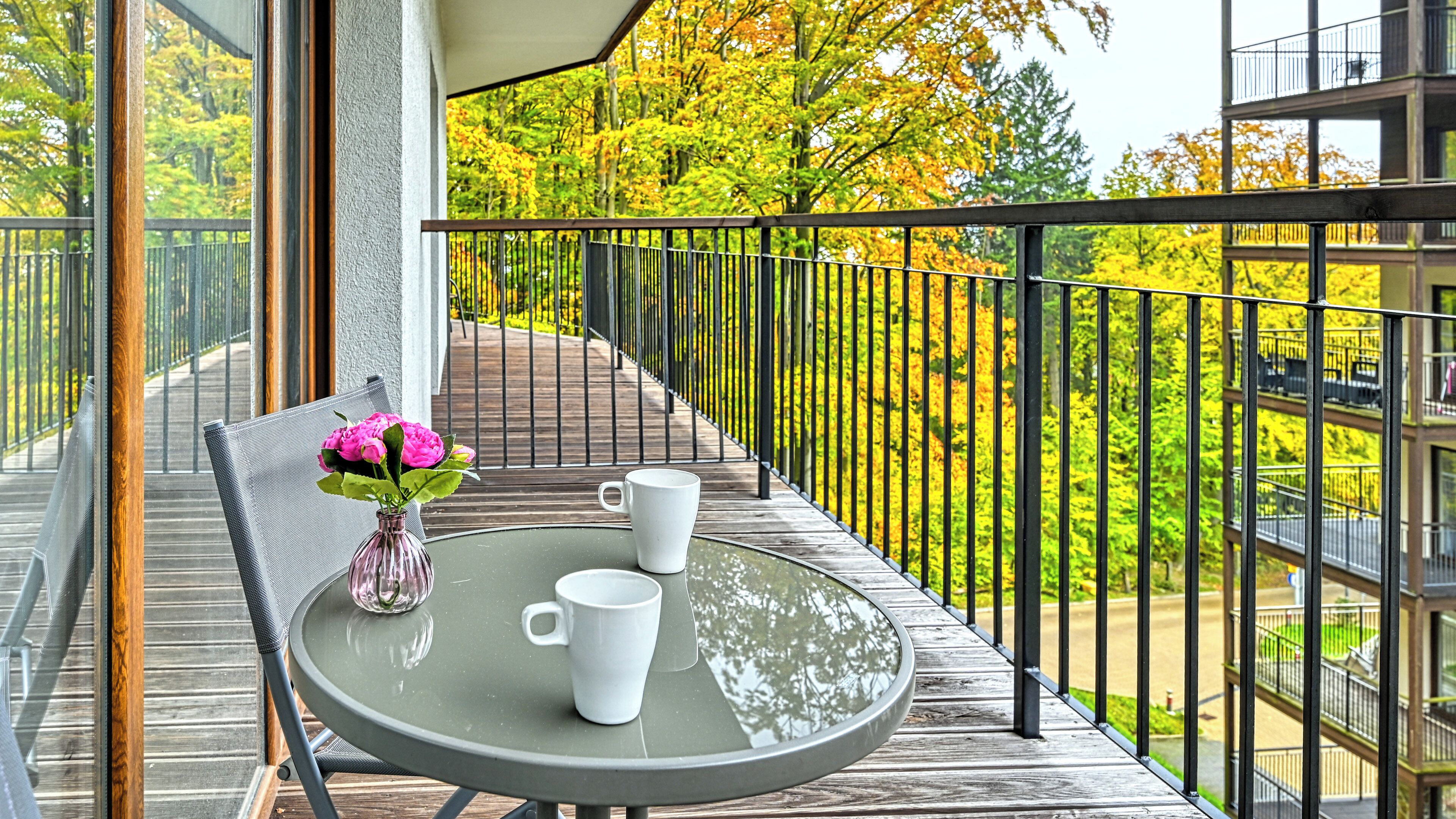A balcony with a round table, two white cups, and a vase with pink flowers.