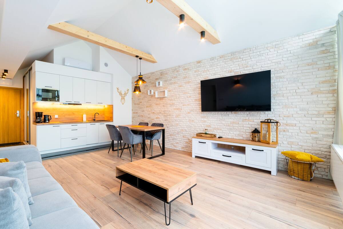 Una sala de estar moderna con sofá, mesa de centro y televisor. Se observa una cocina con gabinetes blancos y pared de ladrillo.