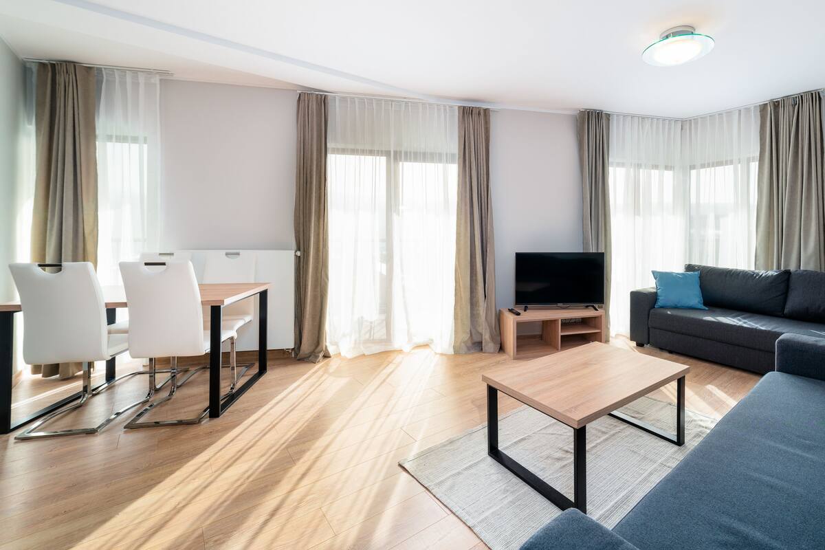 Una sala moderna con un sofá, un televisor, una mesa de comedor con sillas y ventanas grandes con cortinas.