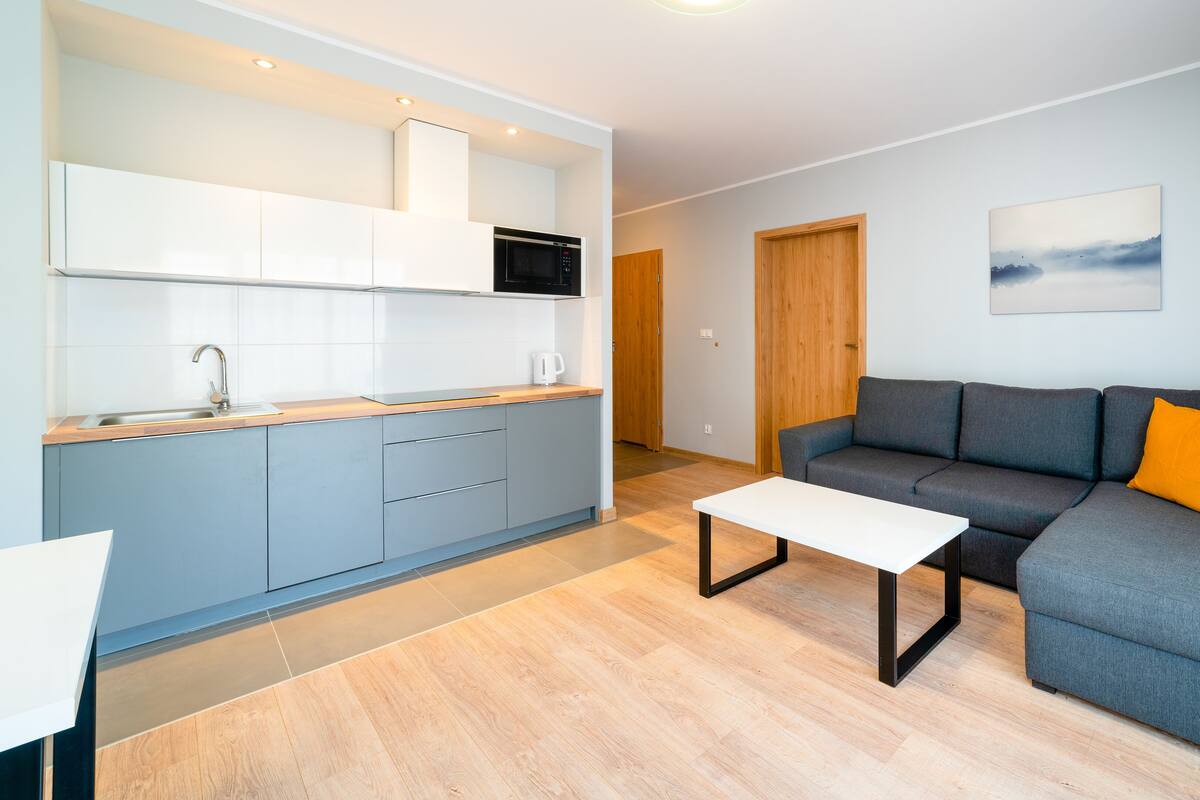 Una sala de estar moderna con un sofá gris, una mesita de centro blanca y una cocina con armarios blancos y un microondas.