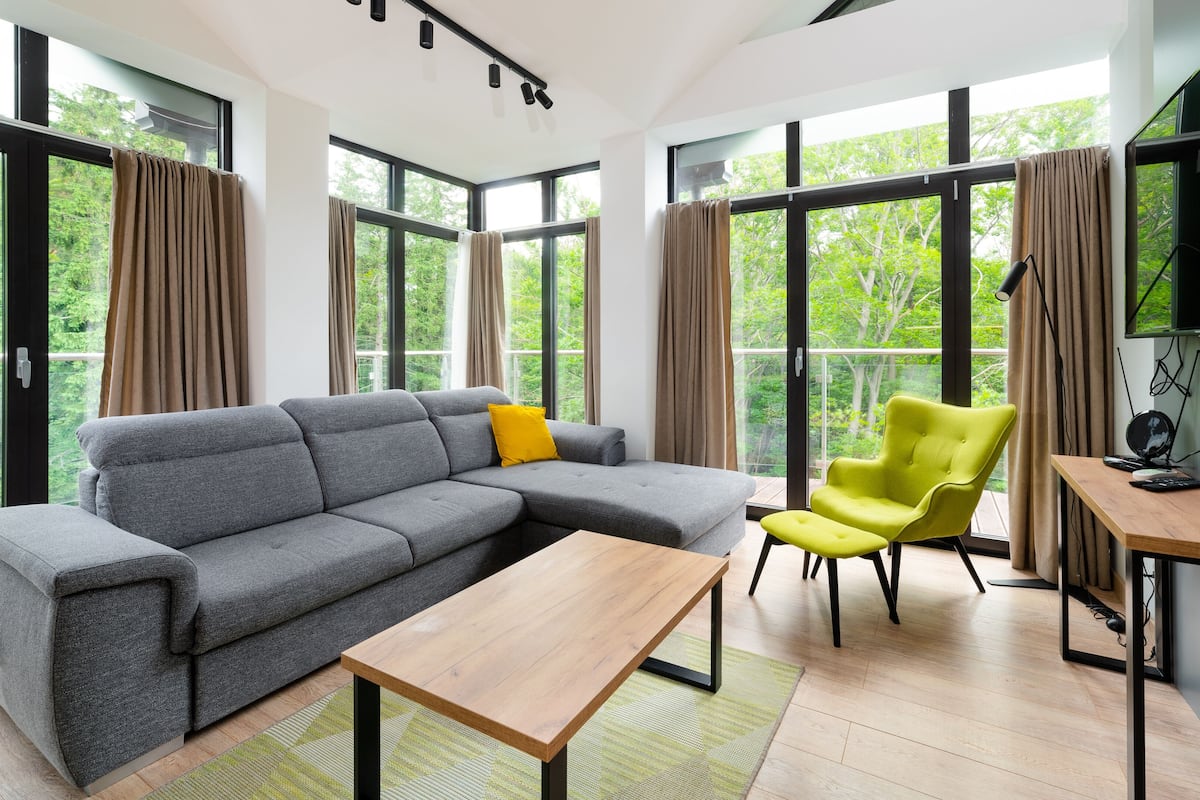 Una sala moderna con un sofá seccional gris, un sillón amarillo, una mesa de centro de madera y una mesita auxiliar con una lámpara. Amplias ventanas ofrecen vistas a zonas verdes.