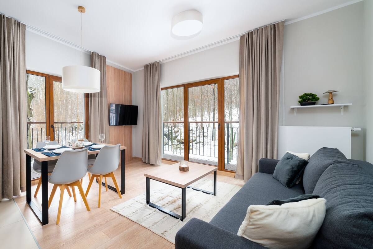 Un salón moderno con un sofá, una mesa de comedor con sillas, un televisor montado en la pared y ventanas grandes con cortinas.