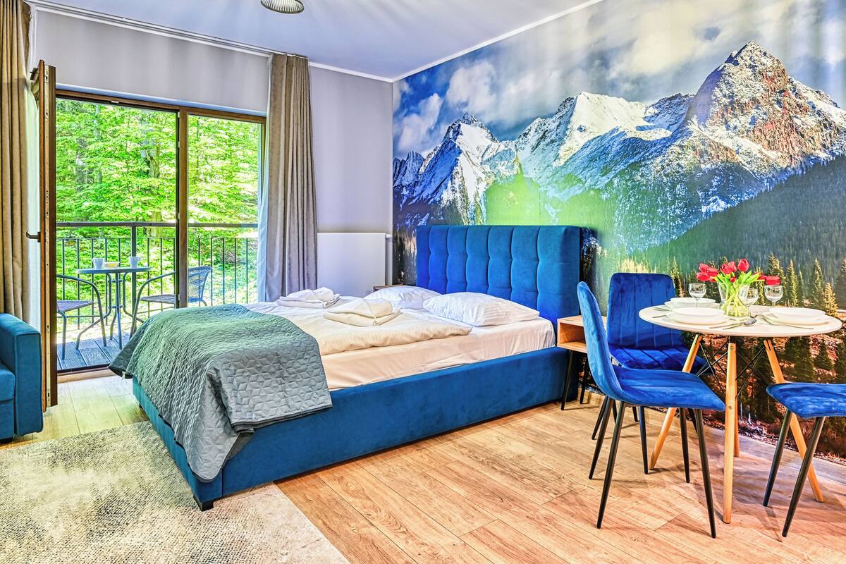 Un dormitorio con una cama grande, una zona de comedor con sillas azules y un mural de montañas.