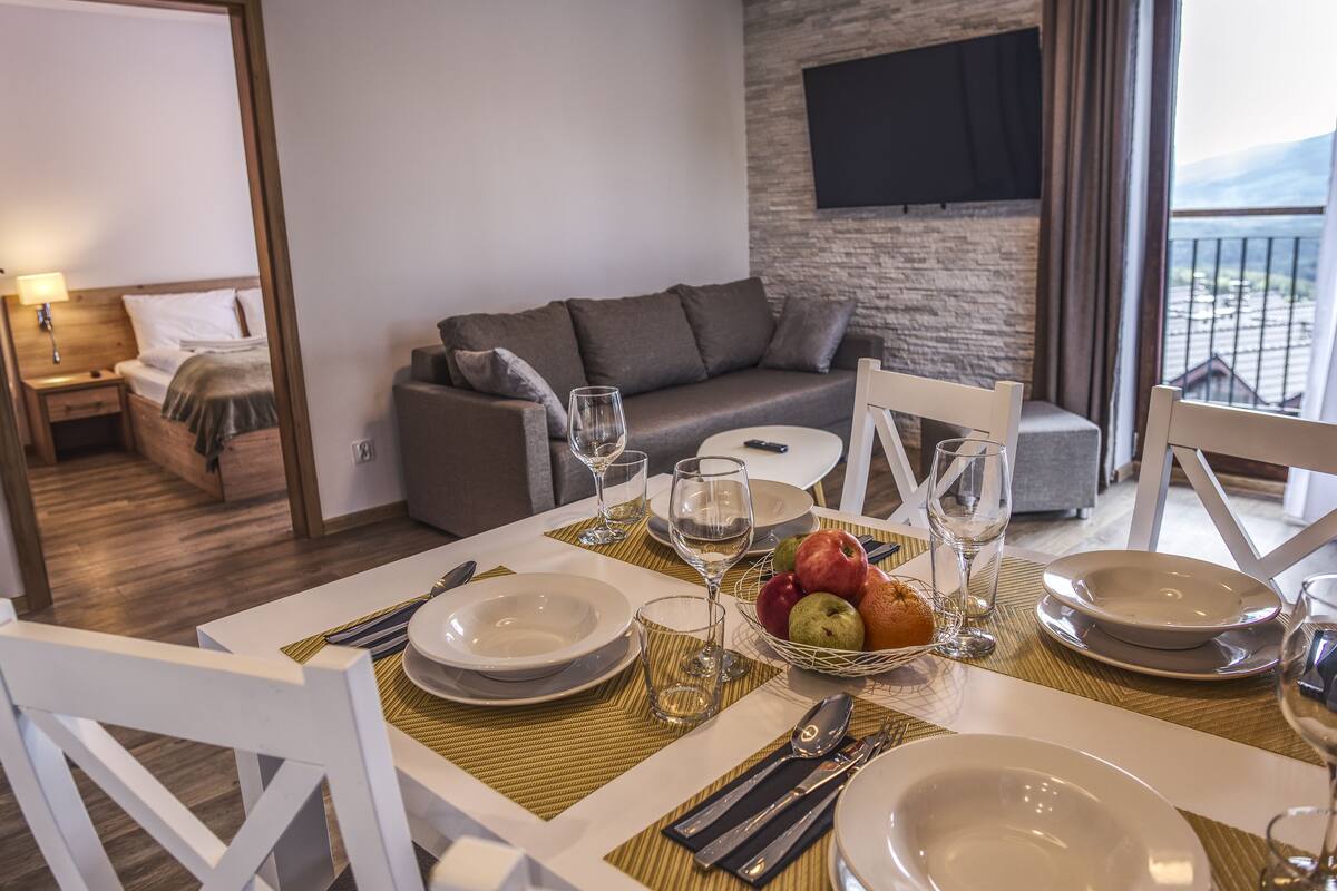 Un comedor con una mesa dispuesta para cuatro personas, incluyendo platos, cubiertos y vasos. Un frutero con manzanas y naranjas está sobre la mesa. La habitación tiene un sofá gris, un televisor montado en la pared y un balcón con vistas.