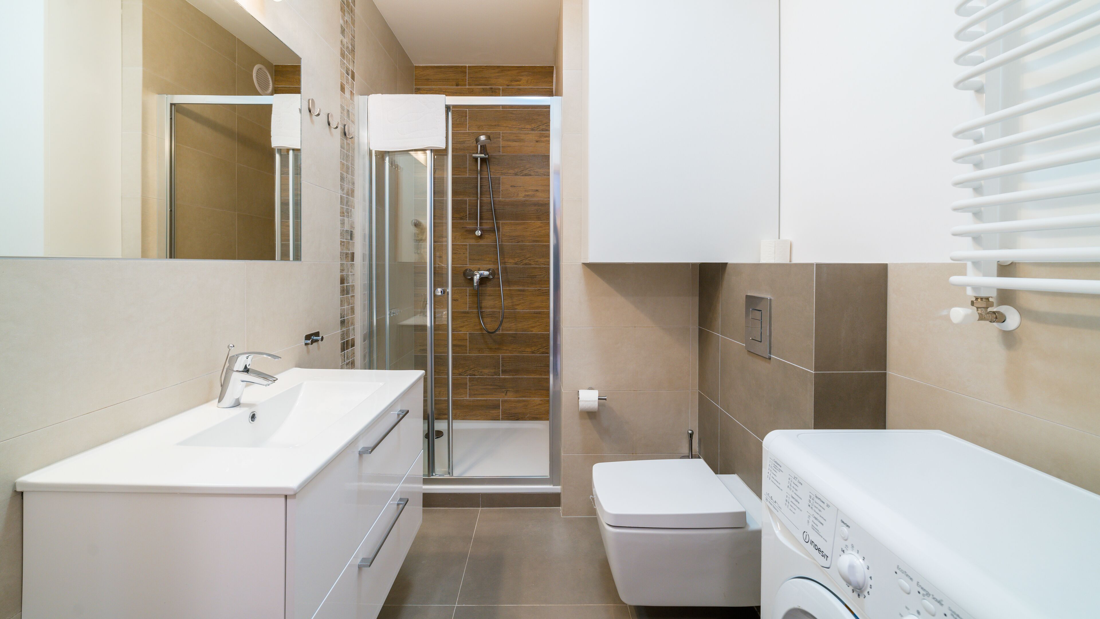Un bagno moderno con lavandino, specchio e servizi igienici bianchi, cabina doccia in vetro e lavatrice a scomparsa.