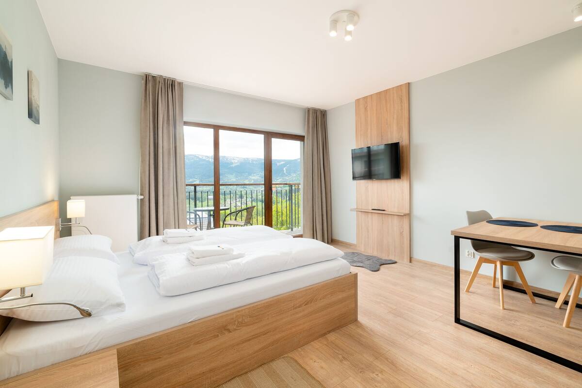Habitación de hotel moderna con una cama grande, un escritorio con dos sillas, un televisor de pantalla plana y un balcón con vistas panorámicas.