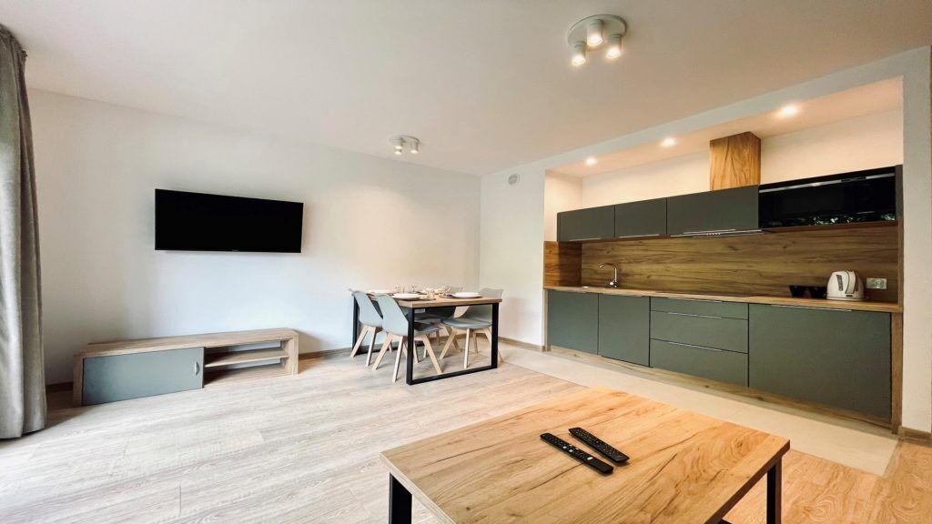 A modern living room with a wooden coffee table, a TV mounted on the wall, and a kitchen area with grey cabinets and a wooden countertop.