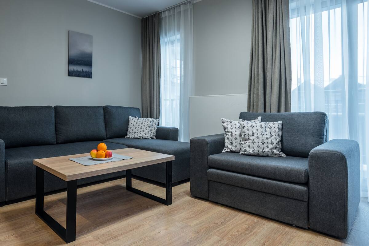 Una sala de estar moderna con un sofá gris, un sillón más pequeño, una mesa de centro de madera con fruta y un cuadro en la pared.