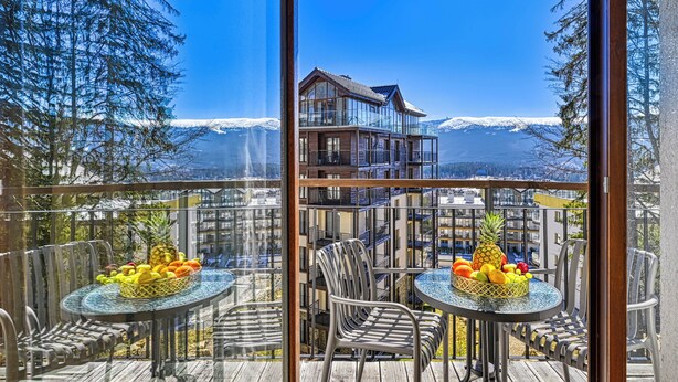 Un balcone con due tavoli, ognuno con un cesto di frutta e sedie, affaccia su un paesaggio montano e un edificio con numerosi balconi.