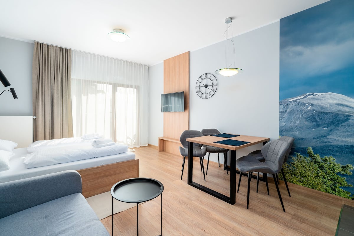 Una habitación de hotel moderna con cama, sofá, mesa de comedor con sillas, televisor montado en la pared y un ventanal con cortinas.