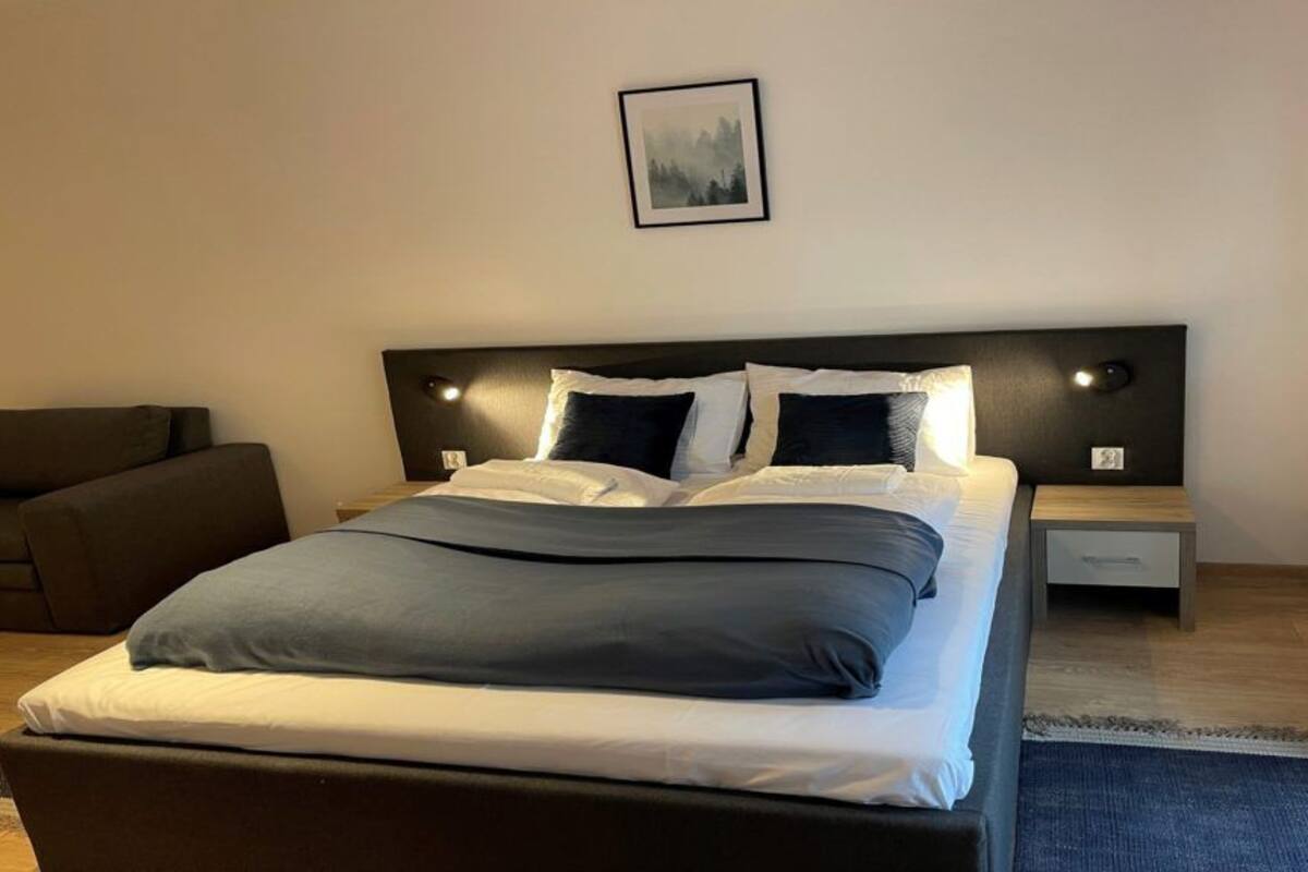 Una cama bien tendida con un cabecero oscuro, una mesita de noche con lámpara y un cuadro enmarcado en la pared.