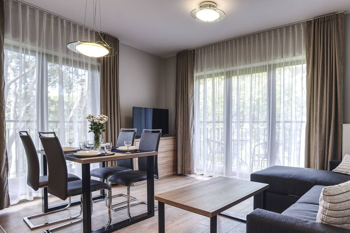 Una sala de estar moderna con un sofá, una mesa de comedor y sillas. Hay un televisor sobre un mueble bajo y una ventana con cortinas.